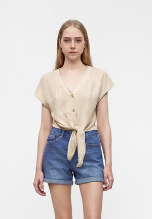 Femme debout devant un fond uni, portant un haut beige à manches courtes boutonné devant, noué à la taille, et un short en jean bleu avec ourlets roulés.