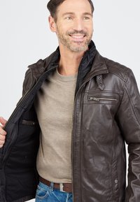 Bruine leren jack met een ritssluiting aan de voorkant, gequiltte details op de schouders en een zwarte binnenvoering. Voorzien van een ritszak op de borst.