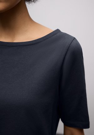 Nærbillede af en person iført en ensfarvet mørkeblå kortærmet skjorte med en bred rund halsudskæring mod en neutral baggrund.