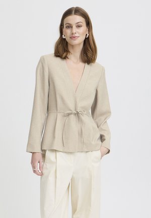Kvinde med skulderlangt brunt hår iført beige omslagsjakke bundet ved taljen og cremefarvede bukser med vide ben, stående foran hvid baggrund.