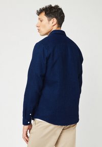 Marineblauwe geweven shirt met een gestructureerde stof, lange mouwen, een knoopsluiting bij de mouwen en kraag, vanuit de achterkant getoond tegen een lichte achtergrond.