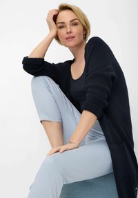 Marineblauwe gebreide cardigan met opgerolde mouwen, gecombineerd met lichtblauwe cropped broek. Model zittend, wat de zachte texturen en de relaxte pasvorm benadrukt.