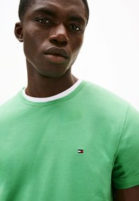 Zielony bawełniany t-shirt z okrągłym dekoltem i białym wykończeniem. Posiada małą naszywkę z logo w kolorach czerwonym, białym i niebieskim na lewej stronie klatki piersiowej.