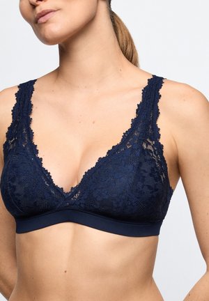 Vrouw draagt een marineblauwe kanten bralette met brede bandjes en bloemmotieven op een lichte achtergrond.