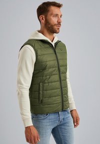 Olivgrön vadderad väst med vertikal sömnad, dragkedjestängning och sidofickor; bärs över en vit hoodie; minimalistisk logotyp på bröstet.