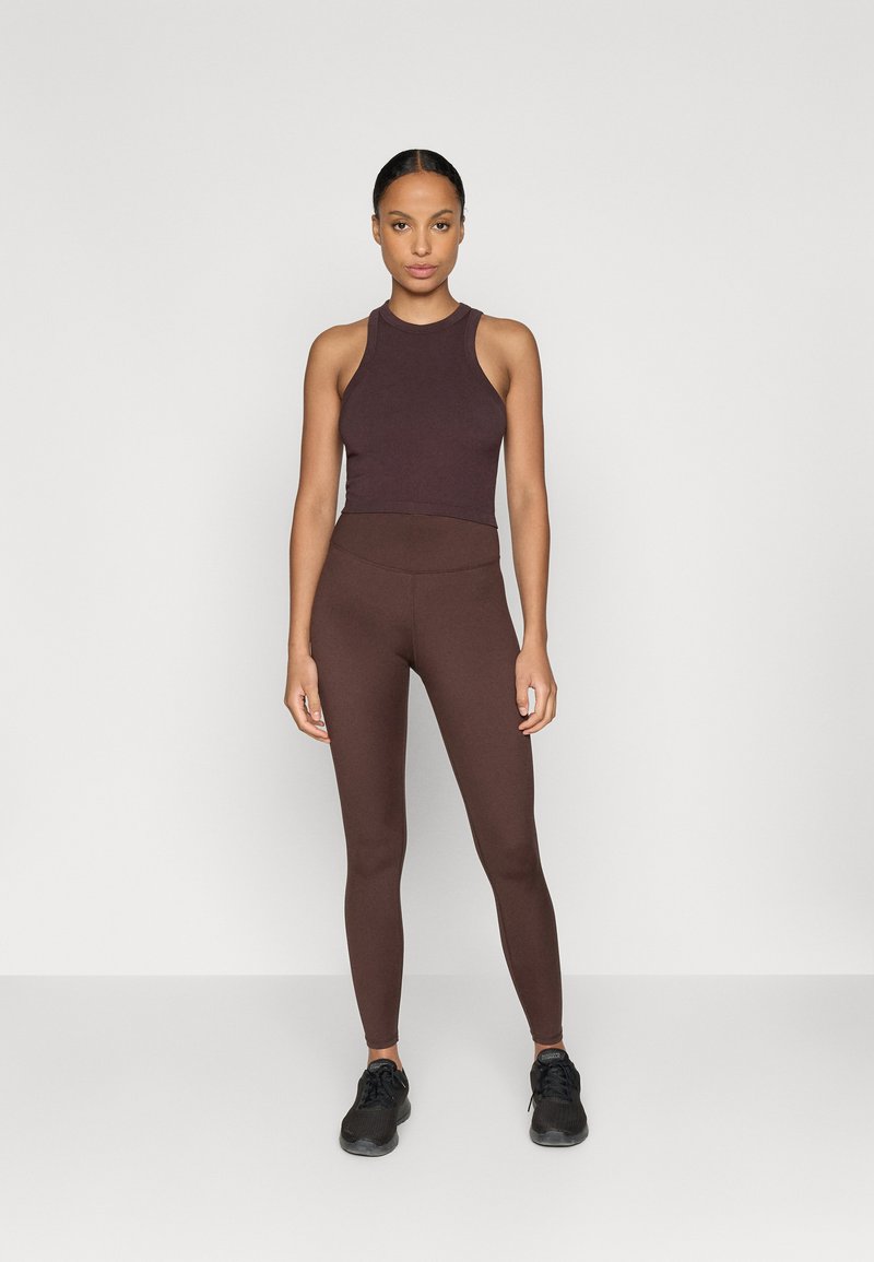 Bruin sportoutfit met een mouwloos topje en hoge leggings. Het materiaal is soepel en heeft een strakke pasvorm. Zwarte sneakers maken de look af.