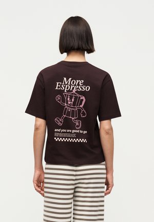 Άτομο με κοντά καστανά μαλλιά φοράει μαύρο T-shirt με ροζ εικόνα ενός περπατούντος μπρίκι και το κείμενο «More Espresso» και μπεζ ριγωτό παντελόνι.