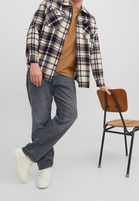 Camisa de cuadros en beige, azul marino y marrón, sobre una camiseta marrón; combinada con vaqueros gris oscuro y zapatillas blancas. Silla de madera cercana.
