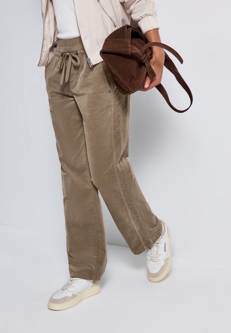 Personne portant un pantalon en velours côtelé beige, des baskets blanches et une veste légère, tenant un sac à main en daim marron sur un fond uni.