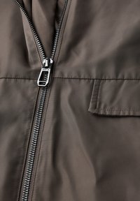 Brun zip-up jacka gjord av slät tyg. Har en silverfärgad dragkedja, en framficka och en något texturerad yta.