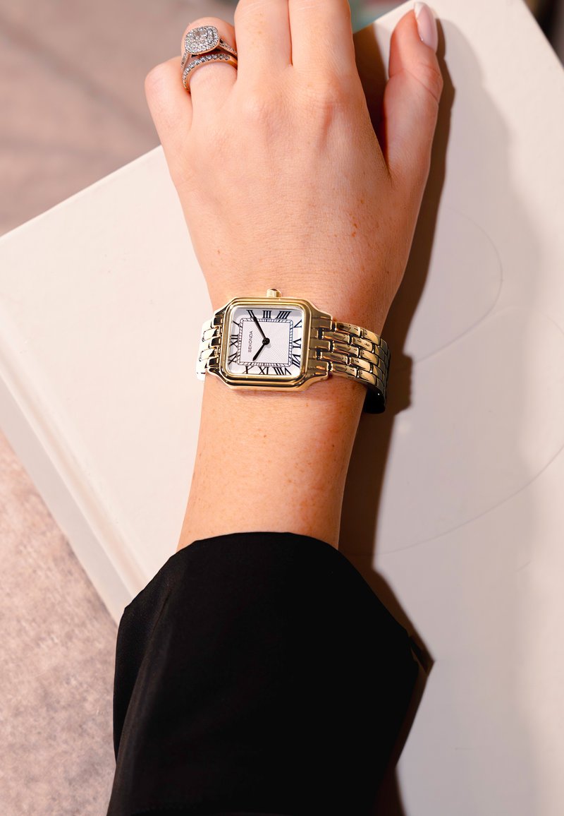 Montre-bracelet dorée avec un cadran rectangulaire, un fond blanc, des chiffres romains et un bracelet en métal maillé, posée sur une main portant une bague.