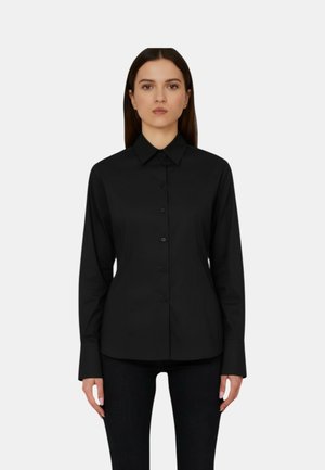 Donna con capelli castani lisci che indossa una camicia nera aderente a maniche lunghe con bottoni e pantaloni neri, in piedi davanti a uno sfondo chiaro e semplice.