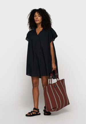 Donna con capelli ricci che indossa un abito nero ampio e sandali neri, che tiene in mano una grande borsa a righe marroni.