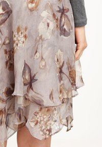 Kukkakuvioinen mekko, jossa on kerroksittain läpikuultavaa kangasta pehmeissä beige- ja ruskeansävyissä. Koristeltu röyhelöreunuksilla ja kevyellä koostumuksella.