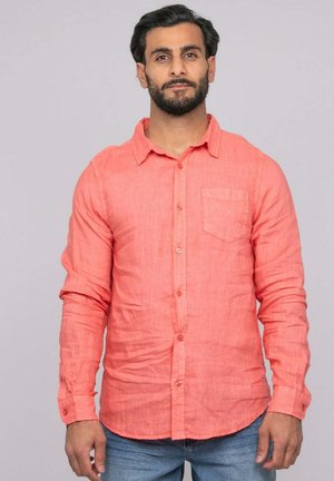 Homme avec barbe et cheveux foncés portant une chemise corail à manches longues boutonnée et un jean bleu, debout devant un fond uni.