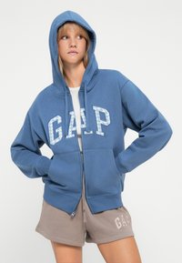 Blå hettegenser med glidelås laget av myk stoff med en stor GAP-logo på forsiden. Har hette og frontlommer. Kombinert med beige shorts.