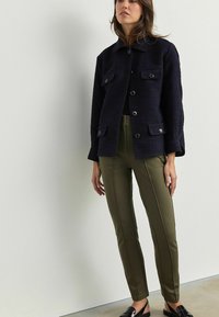 Marineblauwe textuurjacket met knopen en dubbele borstzakken, gecombineerd met olijfgroene slim-fit broek met zijsluitingdetails.