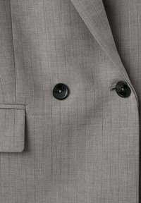 Chaqueta de traje gris a rayas con solapa recta, doble botonadura negra y una tela texturizada con líneas verticales.