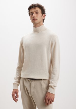 Maglione beige a collo alto in morbido tessuto a maglia, con maniche lunghe e colletto a coste. Abbinato a pantaloni in corduroy di colore chiaro.