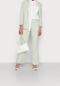 Svetle zelený oblek na míru s širokými kalhotami a sako, doplněný bílou košilí s knoflíky a bílou kabelkou, doplněný bílými sandály na podpatku.