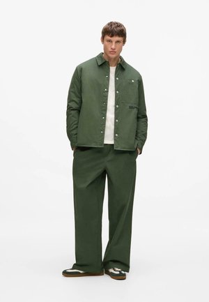 Jeune homme debout, les mains dans les poches, portant une veste verte boutonnée, un pantalon large assorti, un maillot blanc et des baskets.