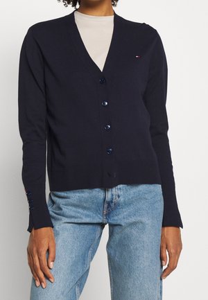 Person iført en marineblå knapcardigan over en lysebeige top og lyseblå jeans, stående mod en ensfarvet baggrund.