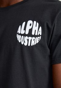 Melnas kokvilnas T-krekls ar izteiktu balto grafisko izdruku "ALPHA INDUSTRIES" aplī, biezā fontā. Īsām piedurknēm.