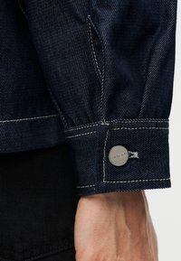 Μανσέτα σκούρου τζην μπουφάν με λευκές ραφές και μεταλλικό κουμπί με χαραγμένο λογότυπο. Το ύφασμα έχει λεία υφή και χαρακτηριστικό διαγώνιο ύφασμα.