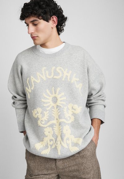 Mand iført en lysegrå sweater med cremefarvet blomsterdesign og teksten "Nanushka", parret med brune strukturerede bukser, hænderne i lommerne, og kigger ned.