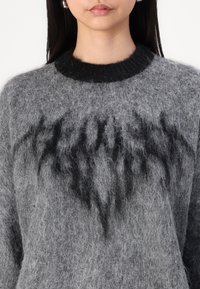 Grijze gebreide trui met een zachte textuur, voorzien van een zwarte ribbelboord en donkere, abstracte patronen aan de voorkant.