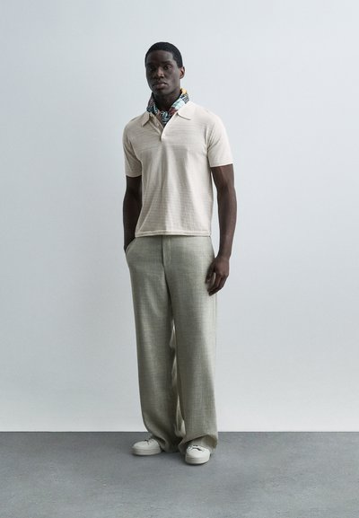 Άνδρας στέκεται μπροστά σε απλό φόντο φορώντας μπεζ κοντομάνικο πουκάμισο, μαντίλι με σχέδιο στο λαιμό, άνετο ανοιχτό γκρι παντελόνι και λευκά αθλητικά παπούτσια.