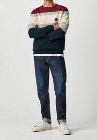 Trui met marineblauwe, crème en bordeauxrode strepen, gebreide textuur, losse pasvorm. Gecombineerd met donkerblauwe spijkerbroek en witte sneakers.