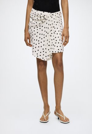Femme portant une jupe blanche mi-longue à pois noirs, un haut noir et des sandales blanches à talons sur un fond uni.
