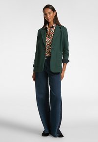 Groen blazer met revers en zakken, gecombineerd met een shirt met patroon en brede spijkerbroeken in een donkere denim. Zwarte schoenen maken de outfit compleet.