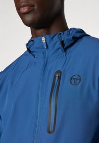 Modrá lehká bunda s kapsou na zip na hrudi, stahovací kapucí a detailem loga na horní levé straně.