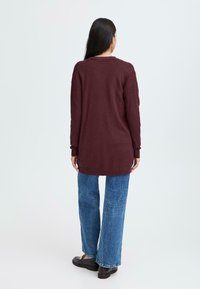 Maglione bordeaux a maniche lunghe con scollo rotondo e polsini a coste, abbinato a jeans in denim blu a gamba larga e calzature scure.