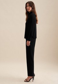 Zwarte blouse met lange mouwen in een ontspannen pasvorm, gecombineerd met op maat gemaakte zwarte broeken. Soepele stof, minimalistisch ontwerp en eenvoudige knoopaccenten.