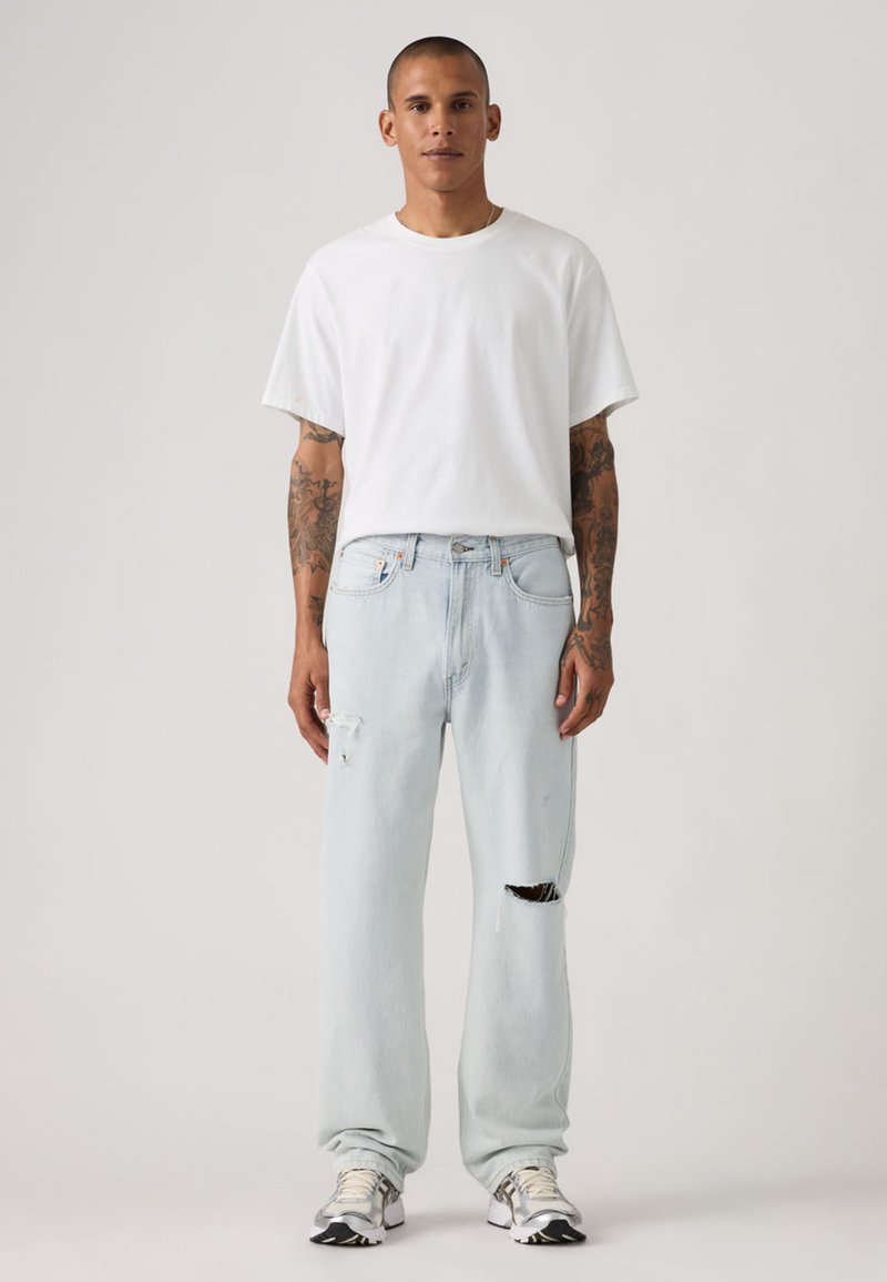 Hellblaue, verwaschene Jeans mit lockerem Schnitt, kombiniert mit einem schlichten weißen T-Shirt. Modell trägt sportliche Schuhe und steht vor einem neutralen Hintergrund.