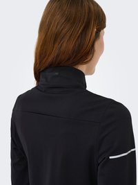 Zwarte top met hoge hals en een gladde textuur, voorzien van een subtiel logo op de kraag en een witte accentenstreep op één mouw.