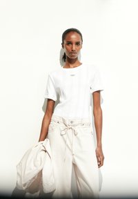 Top blanco de manga corta con un diseño de patrones intrincados, combinado con pantalones sueltos de color beige claro y sosteniendo una chaqueta beige.
