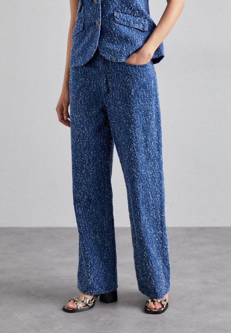 Blue tweed suit pants with a textured finish, wide leg design, and a relaxed fit, paired with black and white patterned sandals.