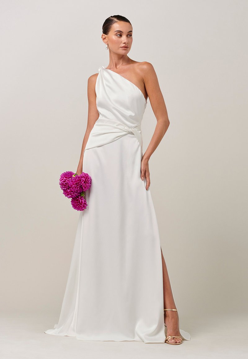 Vrouw in een witte satijnen jurk met één schouder, die een boeket felroze bloemen vasthoudt, draagt gouden sandalen en staat voor een effen achtergrond.