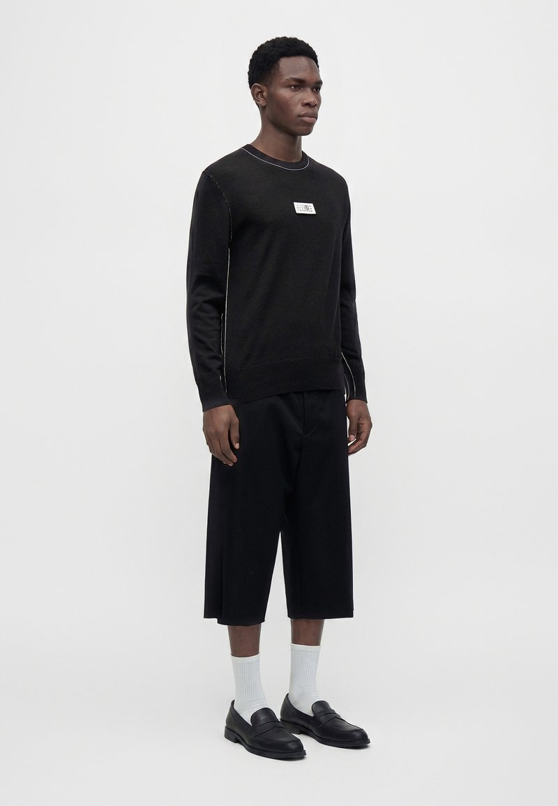 Mann trägt ein schwarzes Langarmshirt mit weißem Etikett, lockere schwarze kurze Hosen, weiße Socken und schwarze Slip-On-Schuhe und steht vor einem einfarbigen Hintergrund.