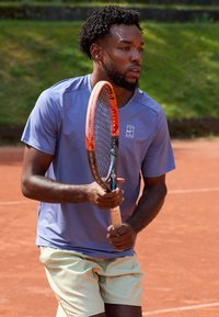 Sinine lühikeste varrukatega sportlik särk, helesinised ruudulised lühikesed püksid, hoides tennisereketit, saviväljakul koos rohelise taustaga.