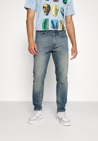 Jeans in denim azzurro chiaro con una vestibilità a gamba dritta, texture sbiadita e design classico a cinque tasche, abbinati a una t-shirt grafica e sneakers.