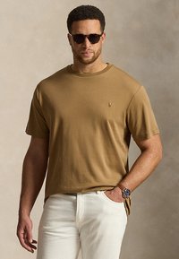 Άνδρας που φορά καφέ μπλουζάκι, άσπρο παντελόνι, σκούρα γυαλιά ηλίου και ρολόι χειρός με το χέρι στην τσέπη, σε ουδέτερο φόντο.