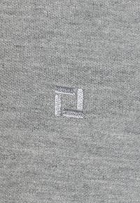 Grauer Stoff mit einer strukturierten Oberfläche, die ein kleines, in silbernem Faden gesticktes Logo prominent auf der Fläche zeigt.