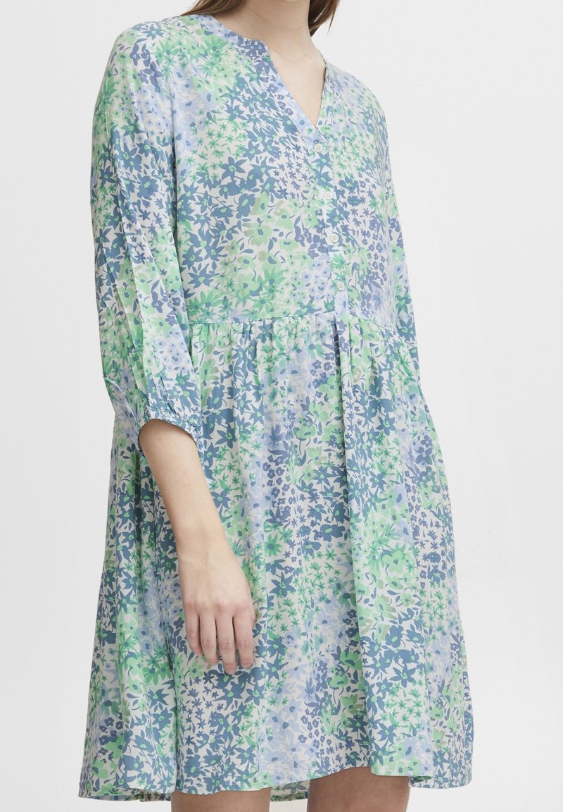 Femme portant une robe à manches longues boutonnée à motifs floraux verts, bleus et blancs sur un fond clair uni.