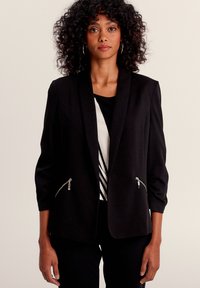 Blazer noir au design ajusté, manches trois-quarts, zips argentés sur les poches et texture lisse. En dessous, une chemise rayée.