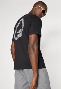 T-shirt noir en coton avec un grand graphique d'une silhouette grise et un logo au dos. Le modèle l'associe avec un short gris et des lunettes de soleil.